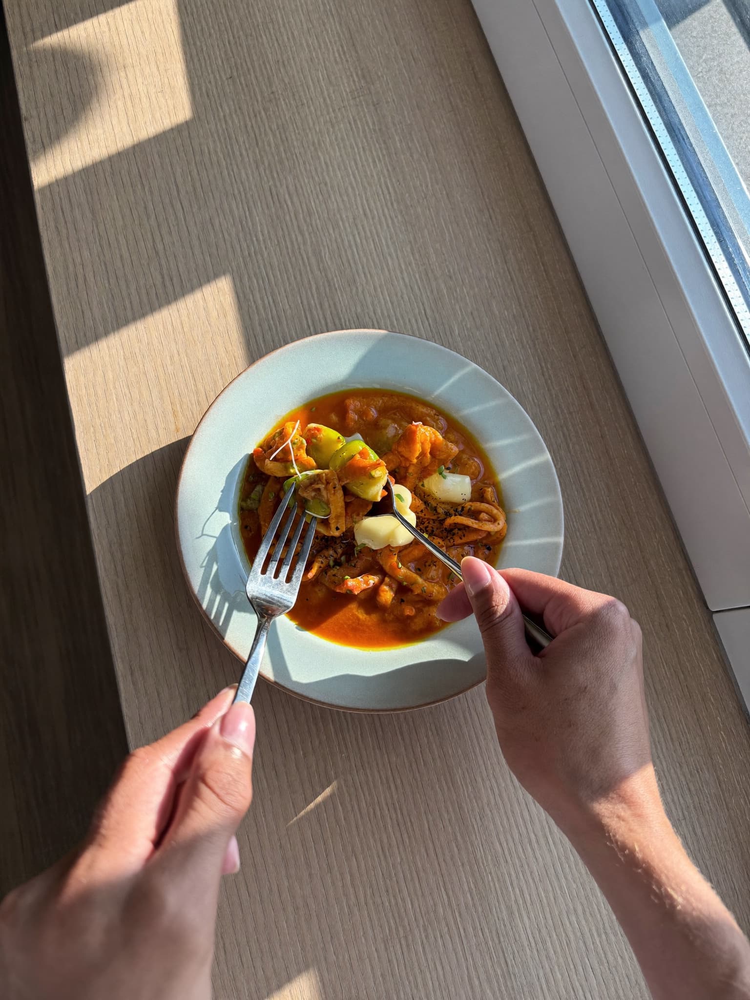 Hands with fork and spoon over a bowl of curry at a sunlit table.