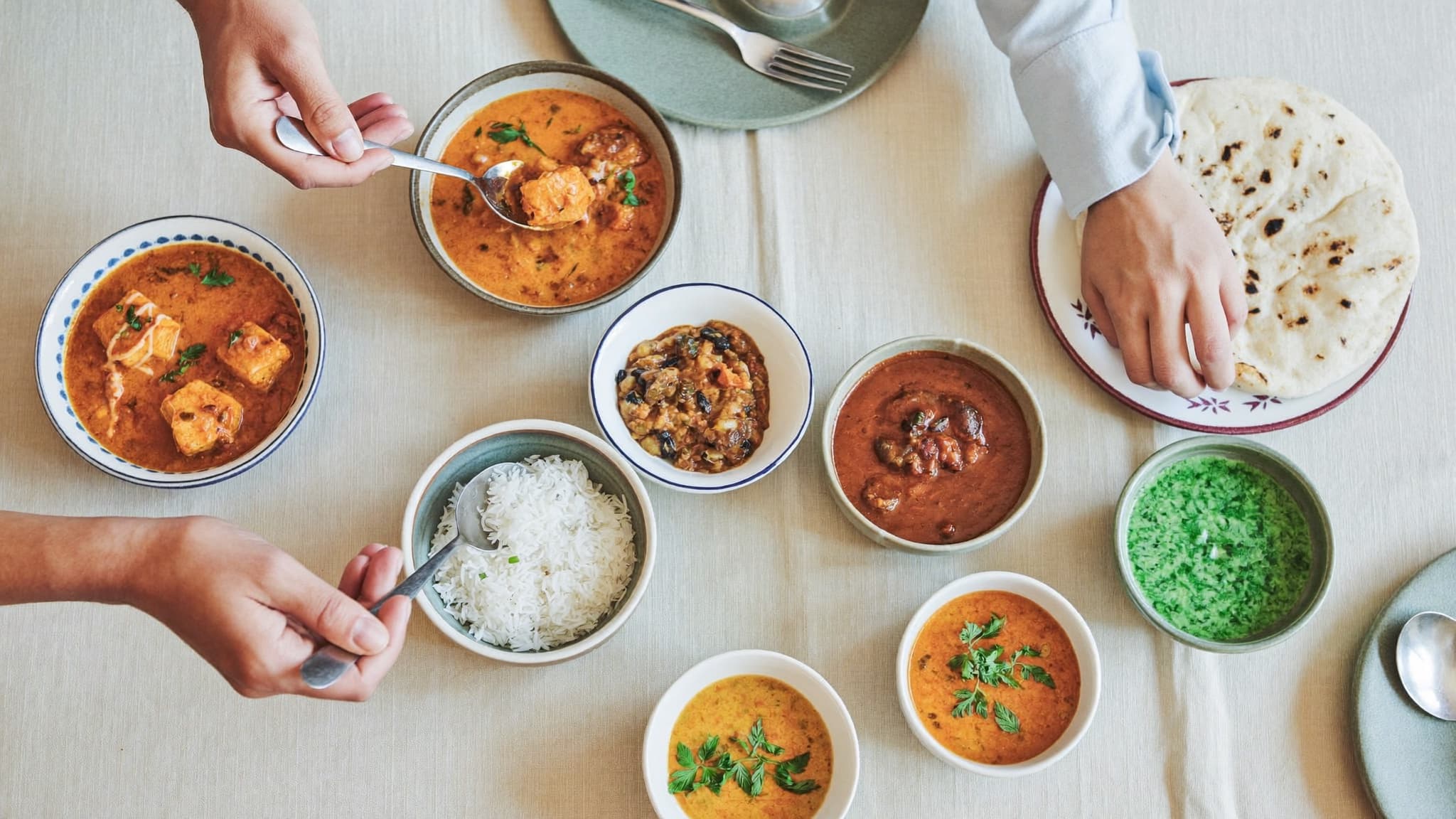 Hands serving curries, rice, and naan family-style across a linen-set table.