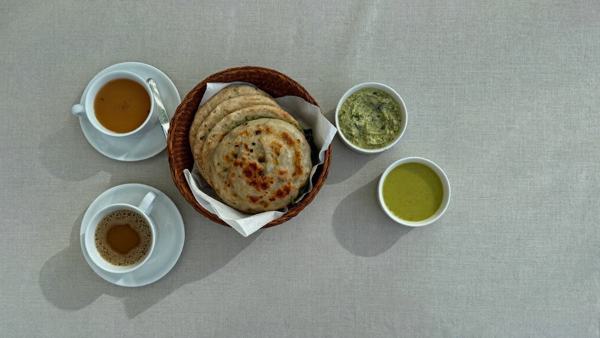 A basket of fresh naan with cups of chai and green chutneys on soft linen.
